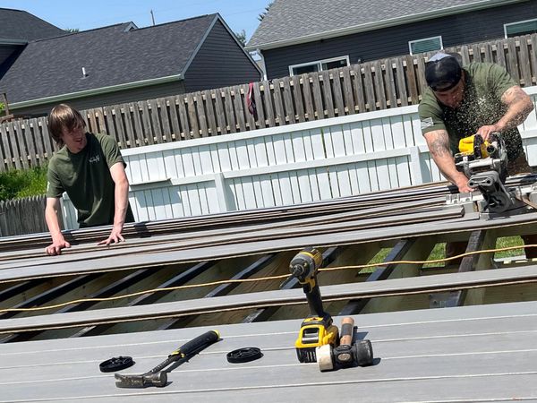 Two people working on a deck outdoors; one uses a saw while the other looks on; tools are nearby.