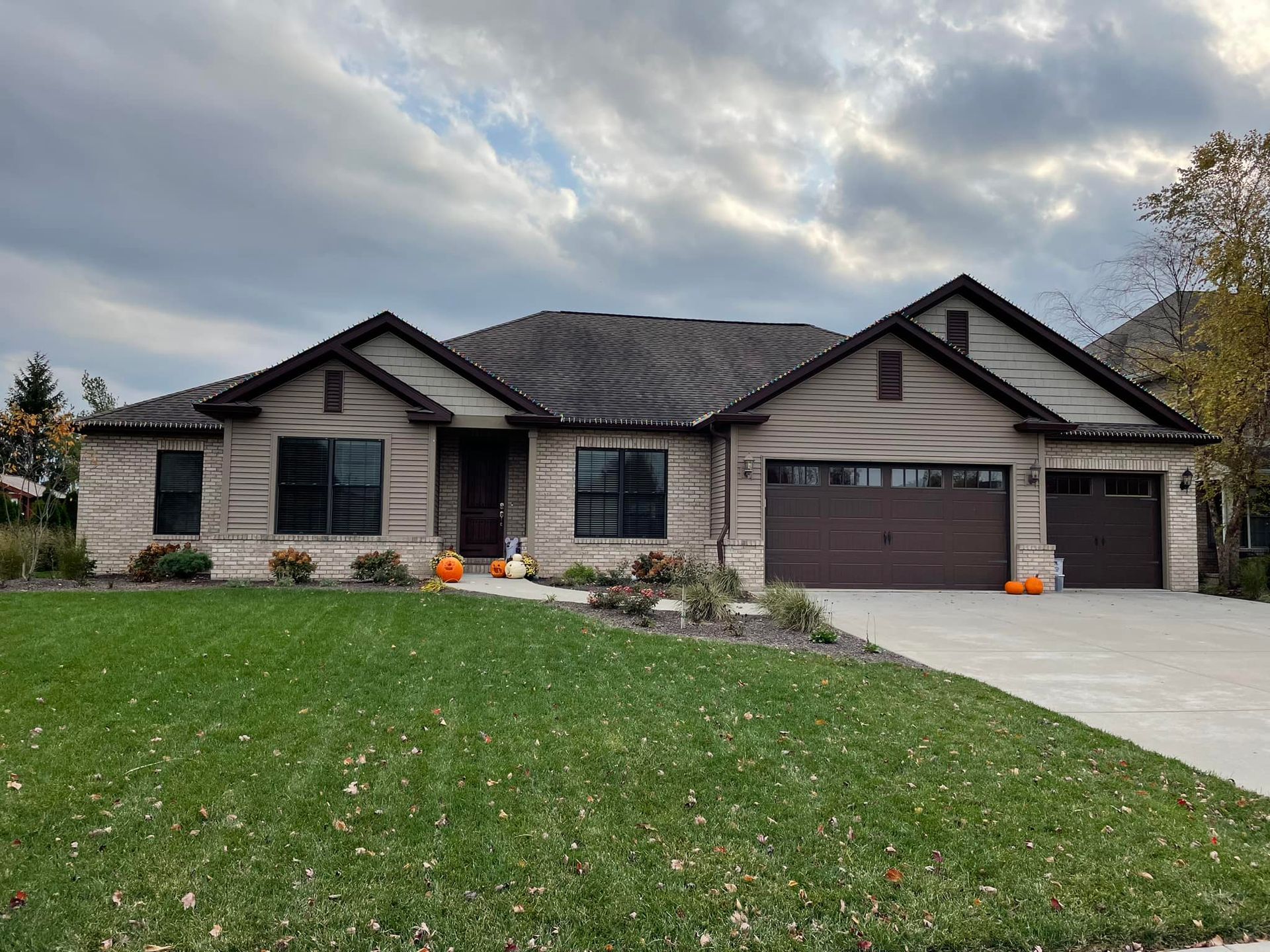 Suburban home with tan brick, brown garage doors, and a green lawn. Two pumpkins sit outside.