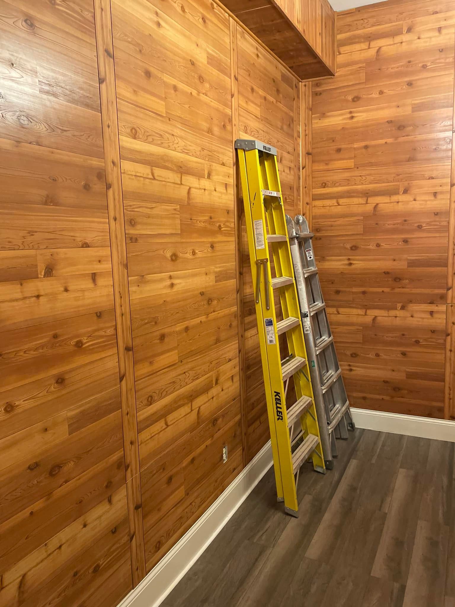 Cedar-paneled walls with two ladders leaning against them, in a room with wood-look flooring.