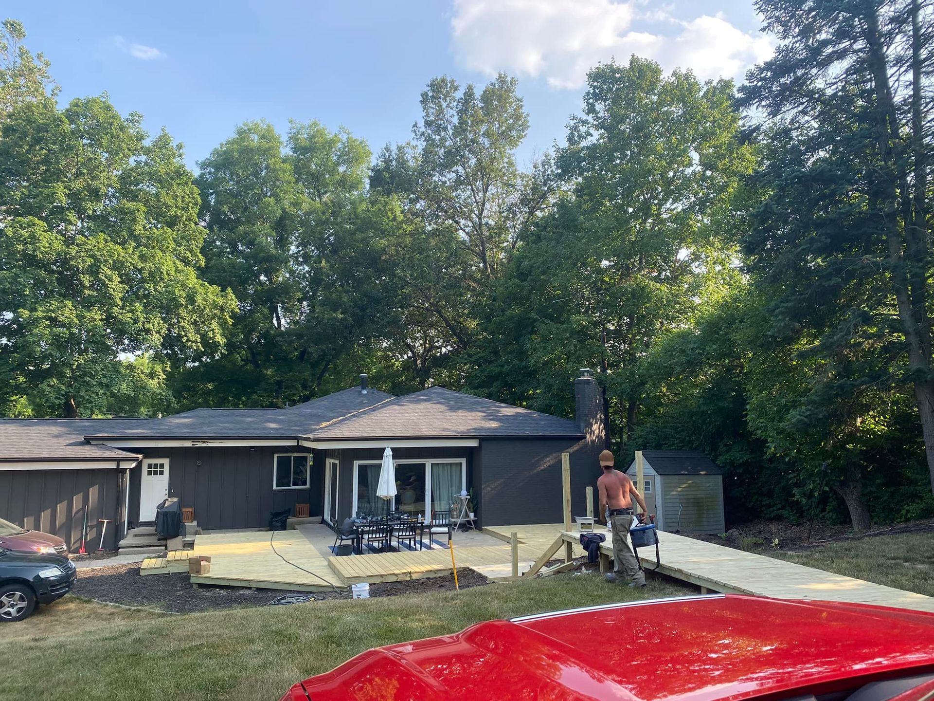 A house with a wooden deck, surrounded by trees; a person works on the deck; red vehicle in foreground.
