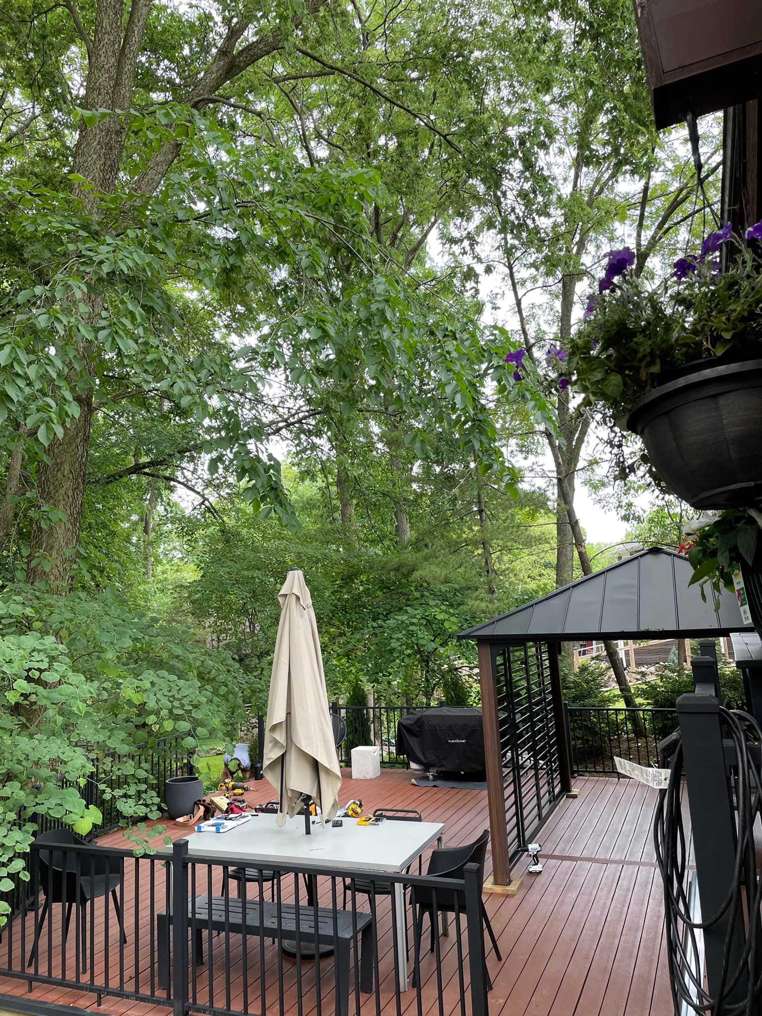 Wooden deck with furniture, umbrella, gazebo, and lush green trees.
