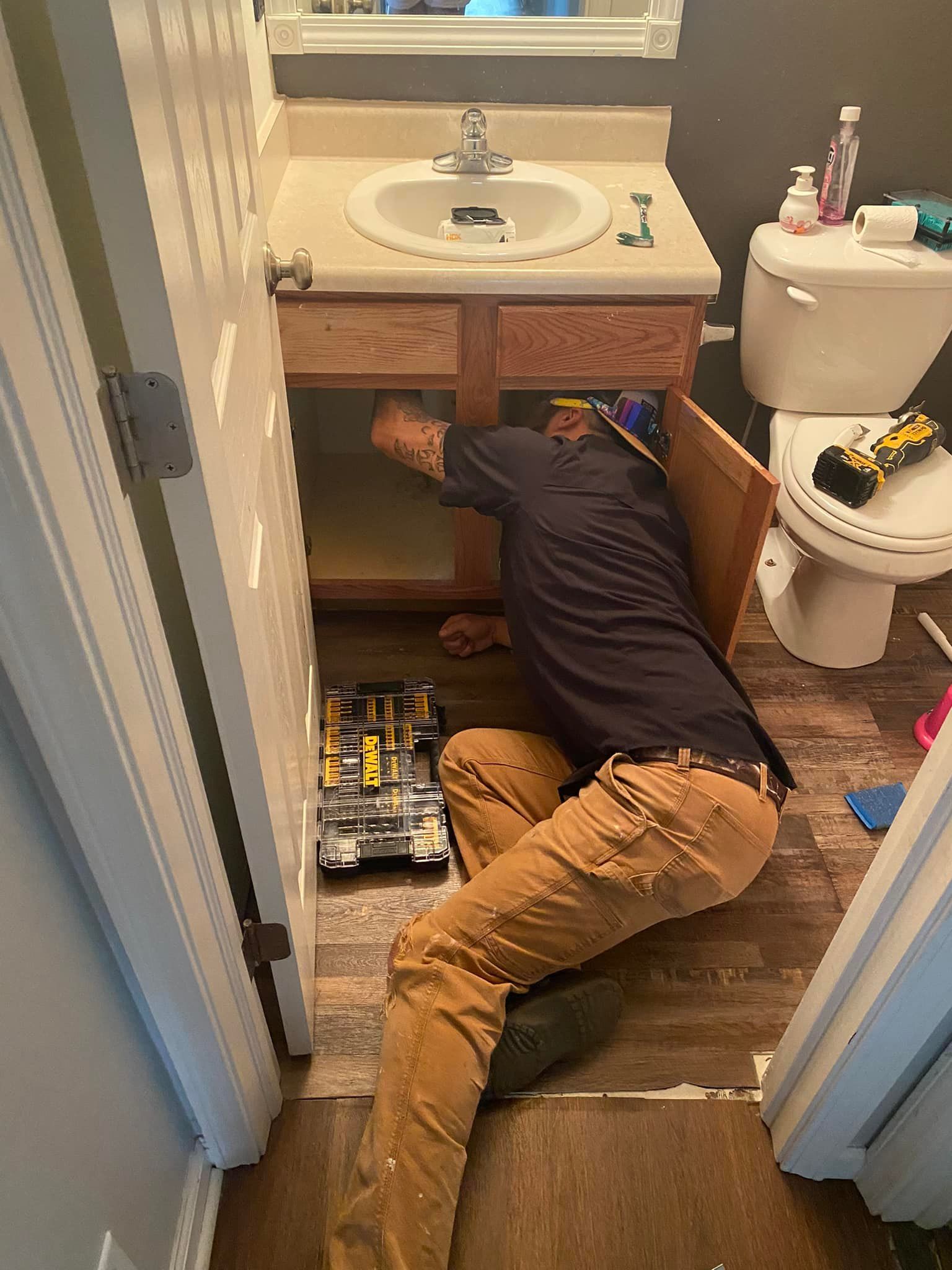 Man working under a bathroom sink, tools nearby. He's on his knees wearing brown pants, fixing something.