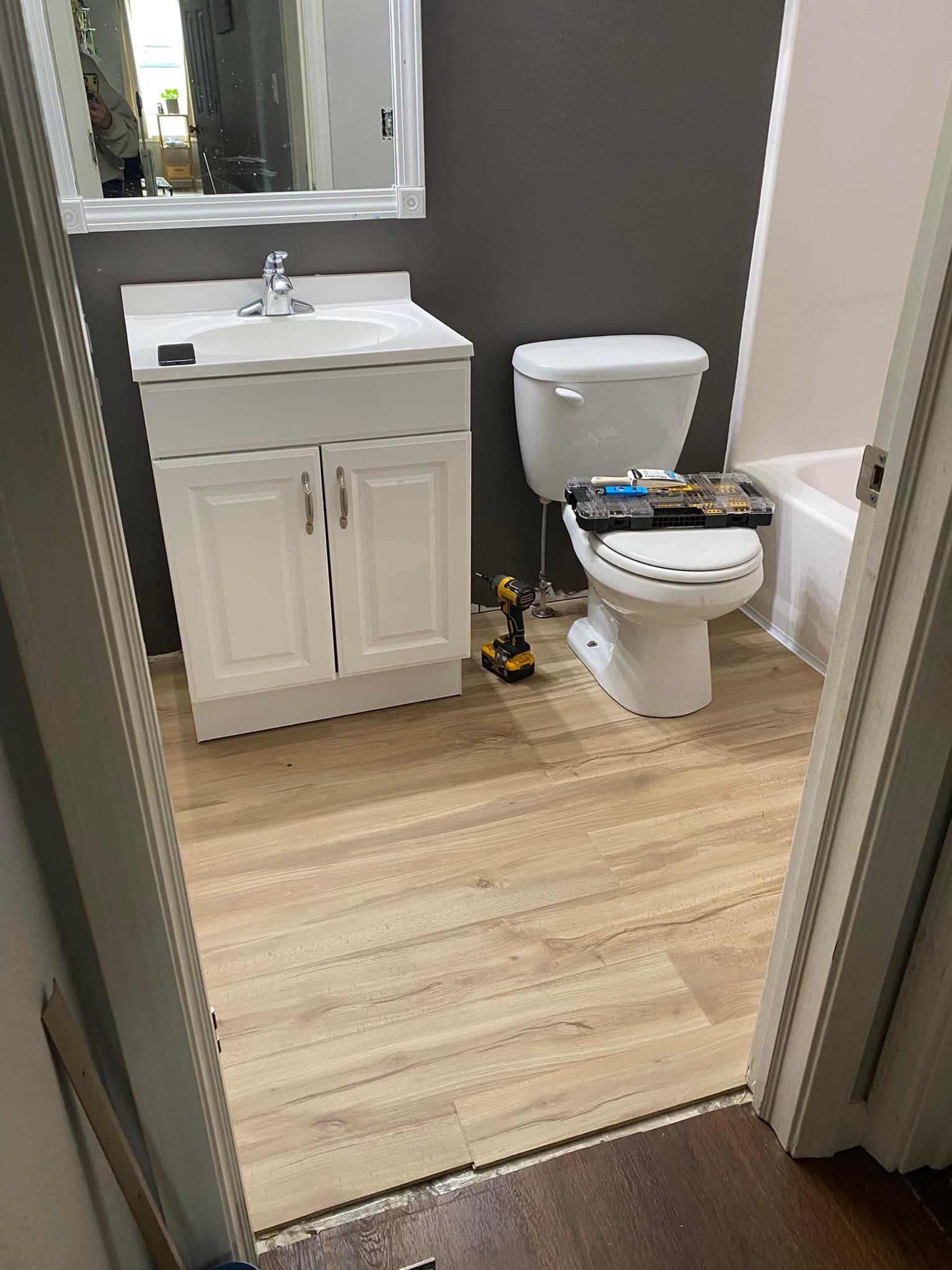 Bathroom with a white vanity and toilet, gray walls, and light wood-look flooring. Tools are on the toilet.