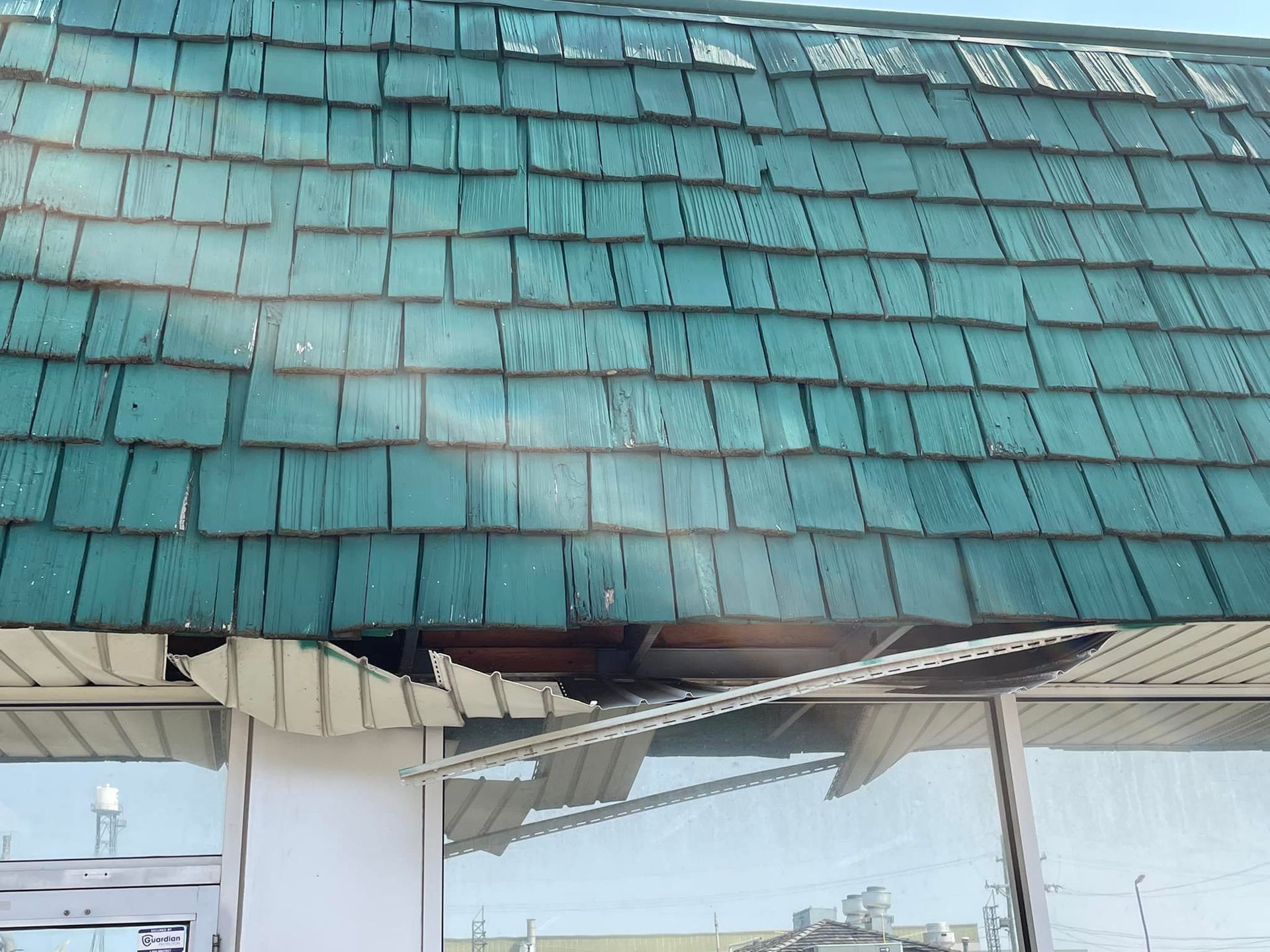 Damaged teal shingle roof over a building with broken panels exposing the interior and glass windows.
