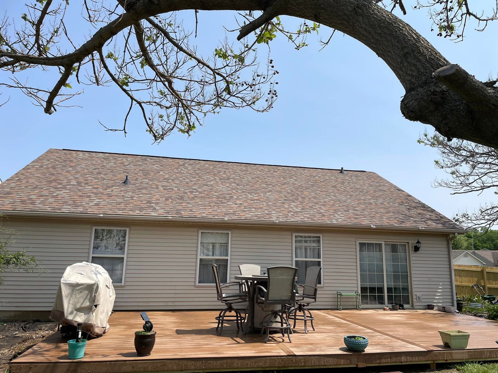 Backyard patio with outdoor furniture, overlooking a beige house under a blue sky, tree branches in the frame.