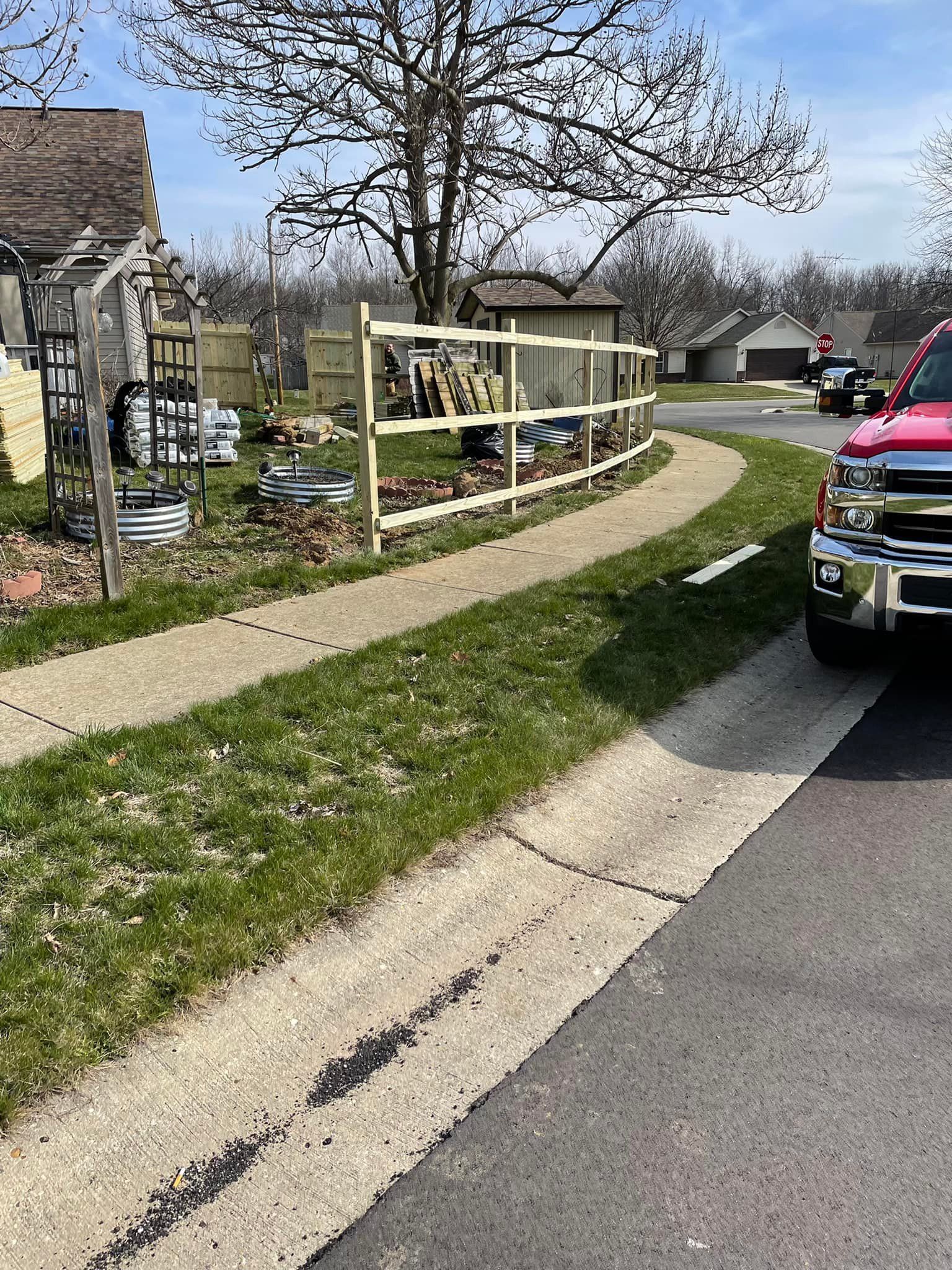 A red truck parked on a street, a wooden fence borders a grassy area next to a driveway.