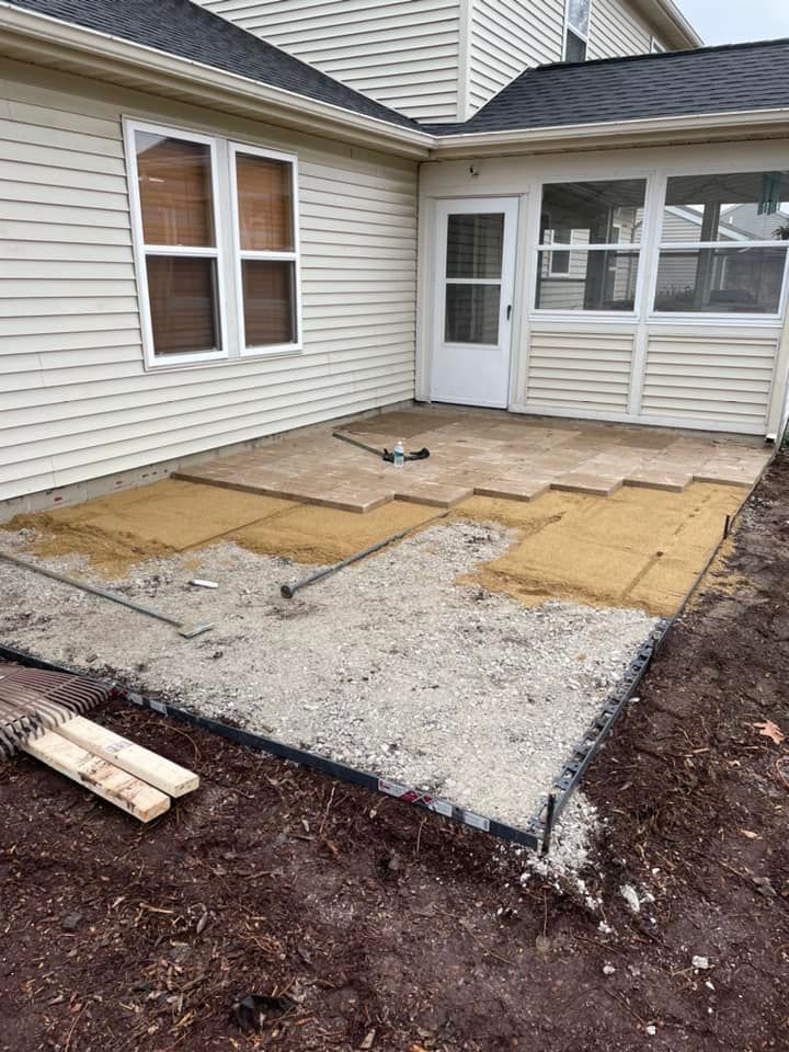 A patio under construction with gravel and pavers. Beige house with windows and door.