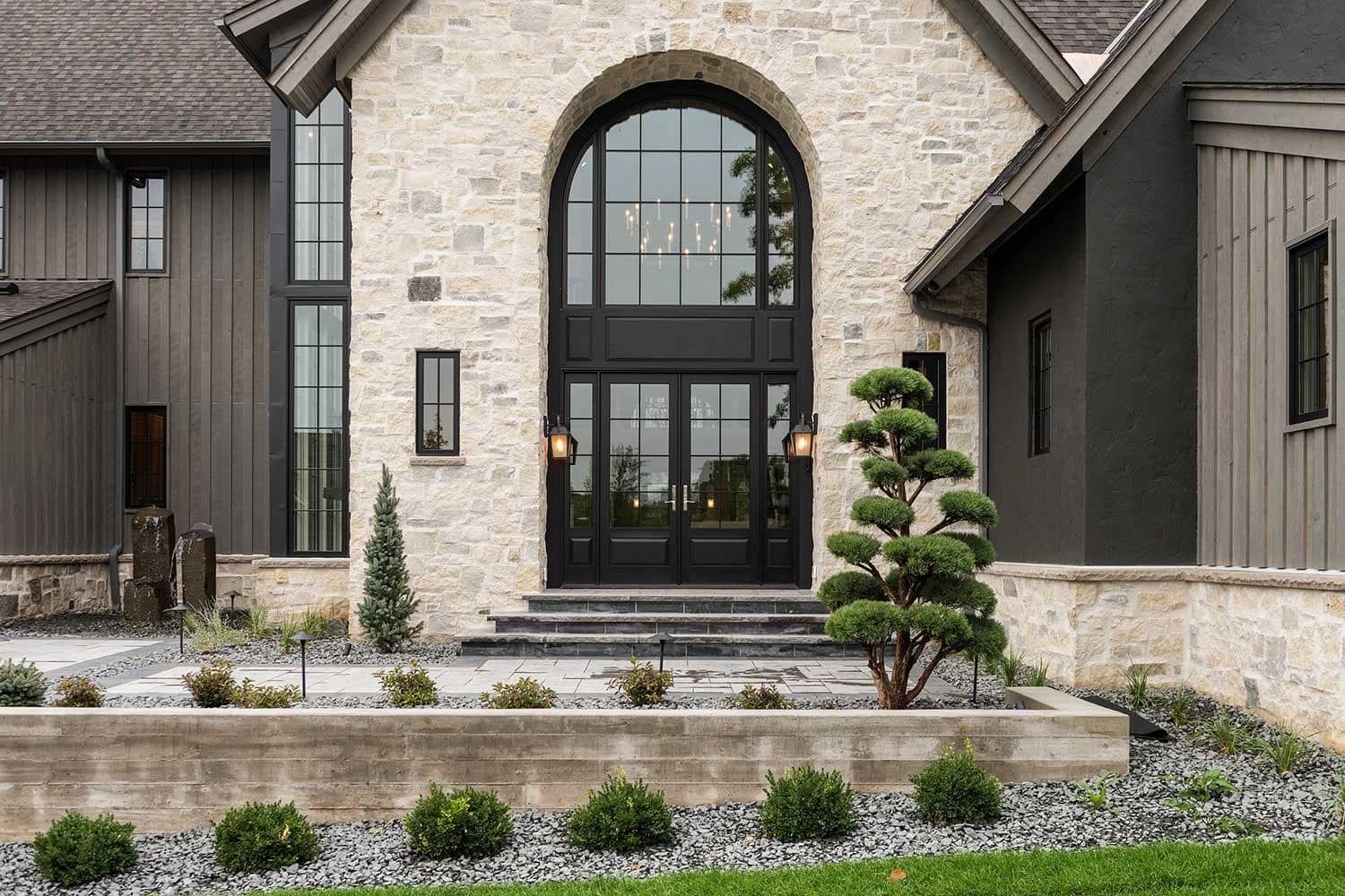 Stone-clad house entrance with arched doorway, black doors, and manicured landscaping.