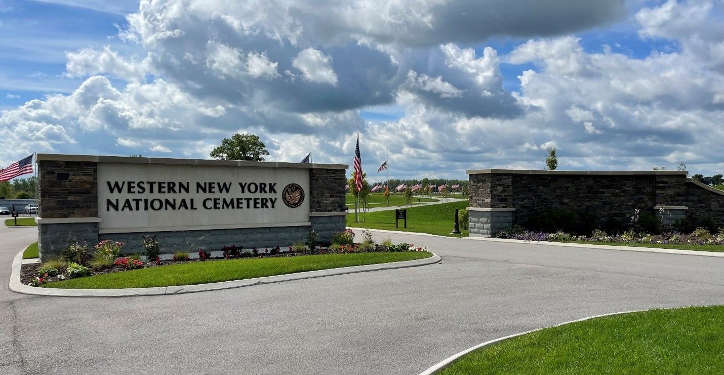 Entrance sign to Western New York National Cemetery with stone pillars and American flags against a cloudy blue sky.