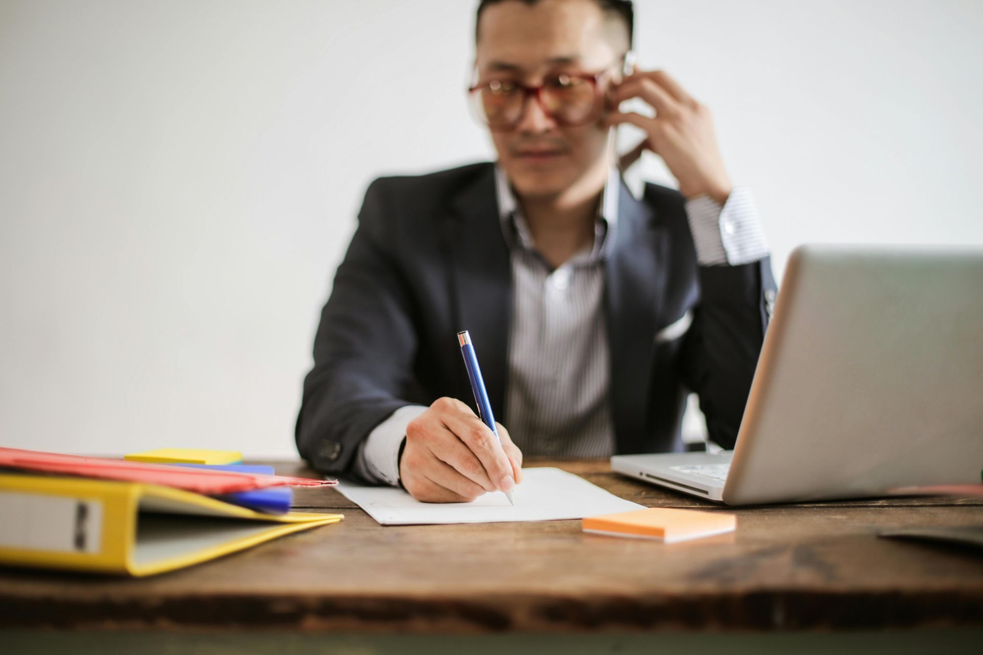 A person in a suit sits at a desk, writing on paper while talking on a phone, with a laptop and yellow folder nearby.