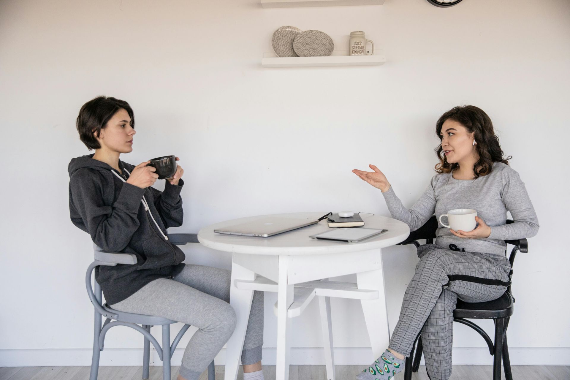 Two people sit at a small white table with laptops and mugs, talking in a brightly lit room with a shelf on the wall.