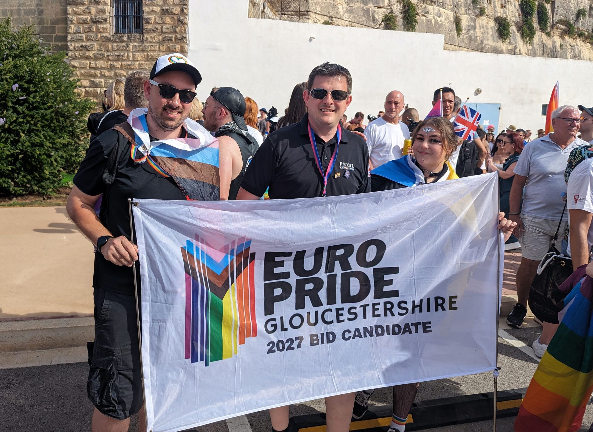 Three happy looking trustees of Pride in Gloucestershire standing behind a white flag promoting EuroPride in Gloucestershire. TEXT: 
