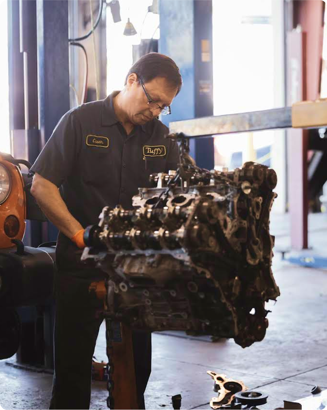 Mechanic examining a car engine in a garage. He is wearing glasses, dark clothes and is focused on the engine.