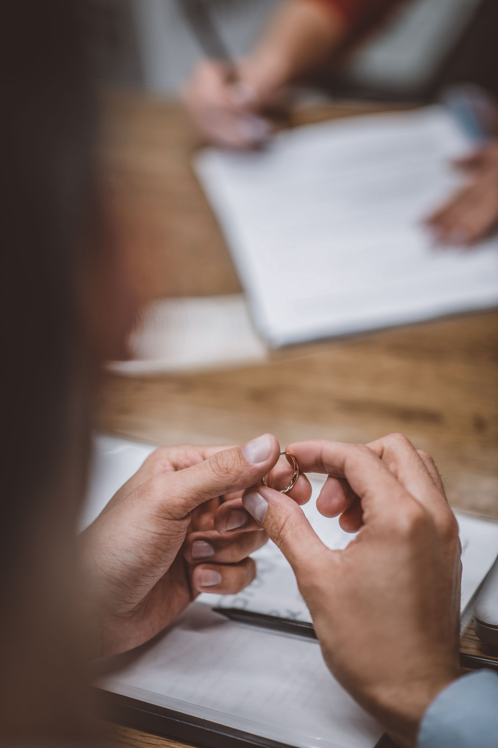 Person holding a wedding ring, possibly in a consultation setting. Others are in the background, with papers on a wooden table.