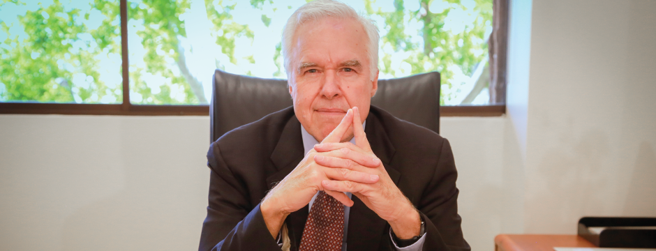 Man in suit, hands clasped, sitting at desk. Background of window with trees.