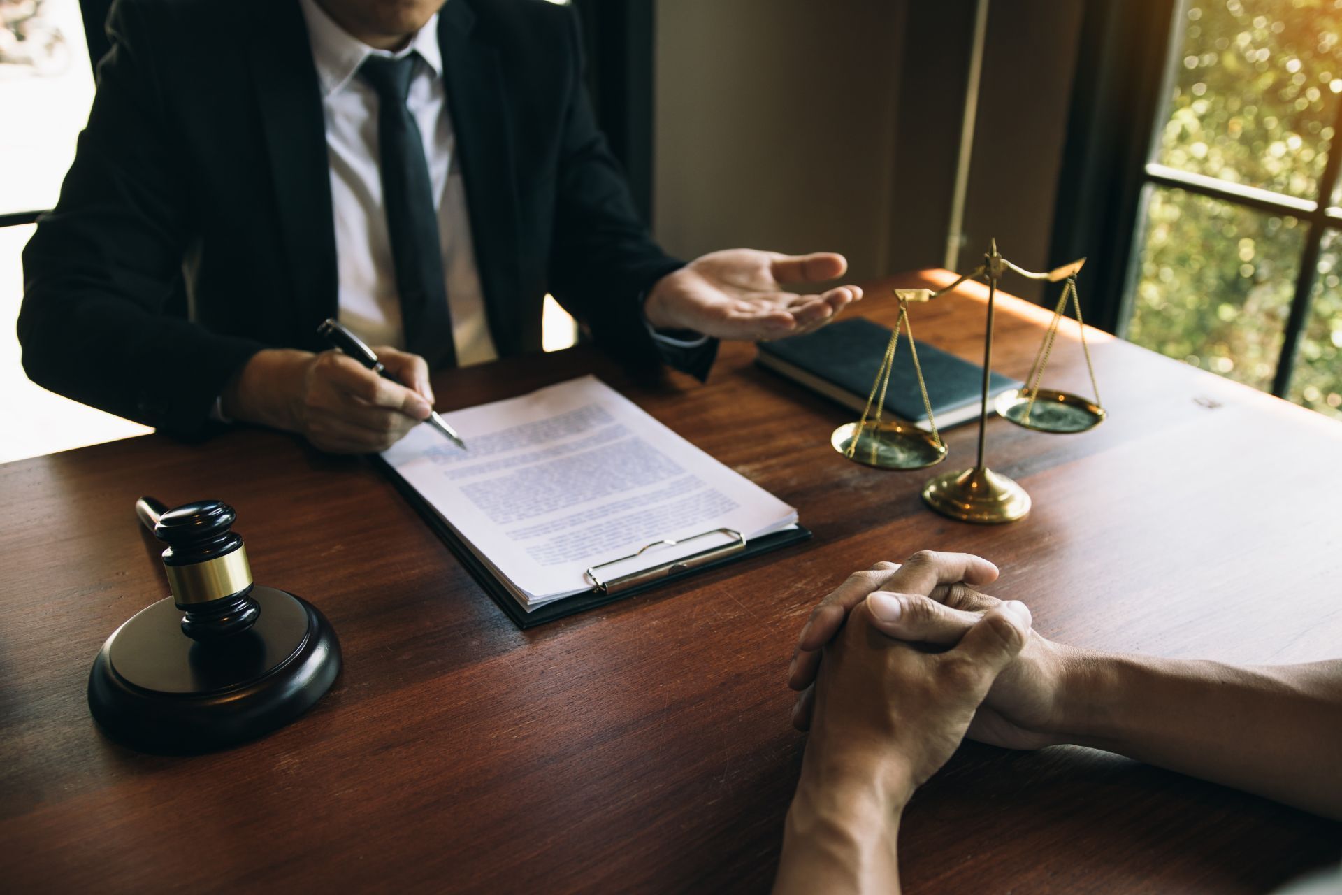 Lawyer in suit gestures to document, client's hands clasped. Gavel and scales on table.