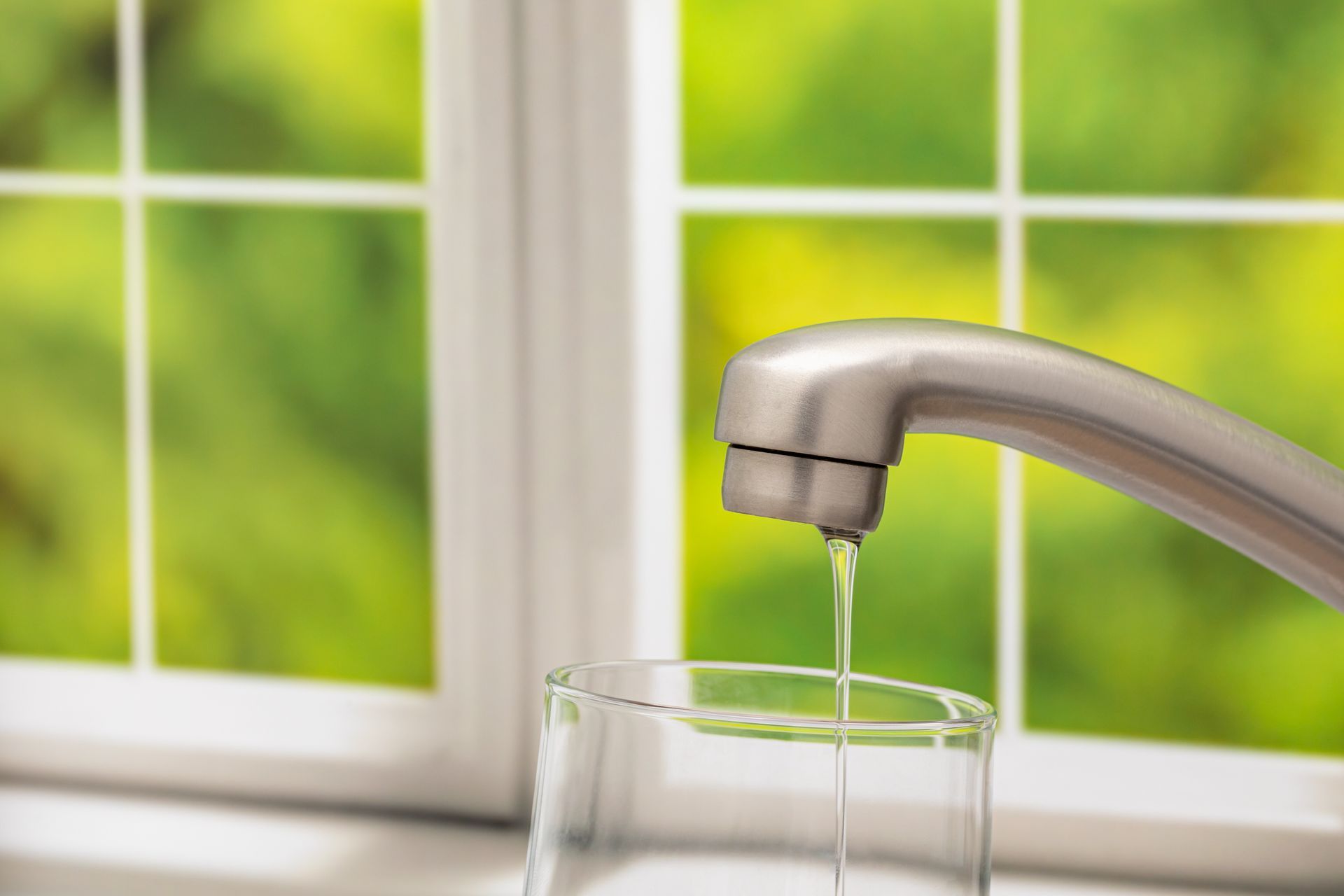 Water pouring from a chrome faucet into a clear glass by a bright window
