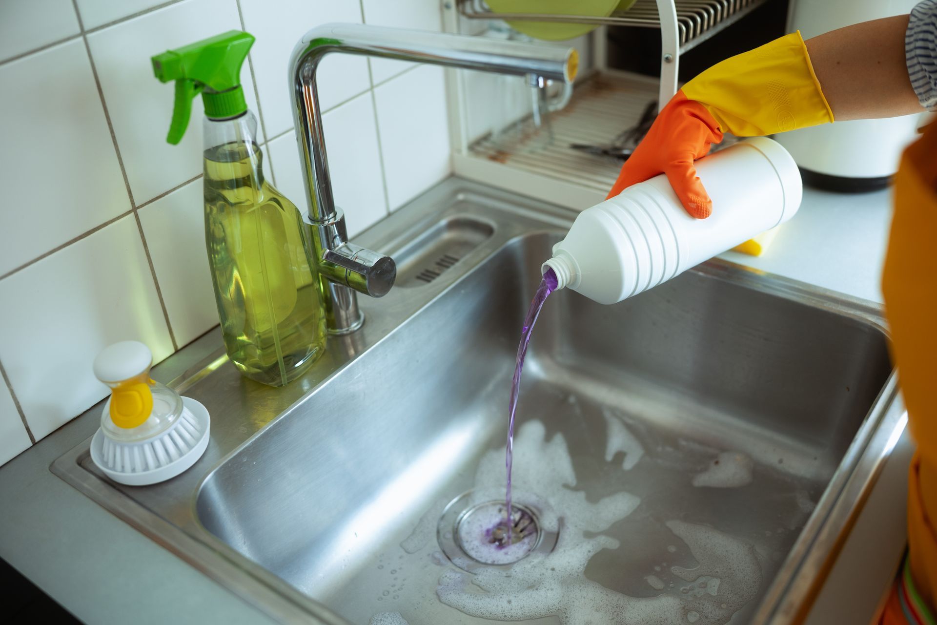 Person in orange glove pours cleaner into a stainless-steel kitchen sink