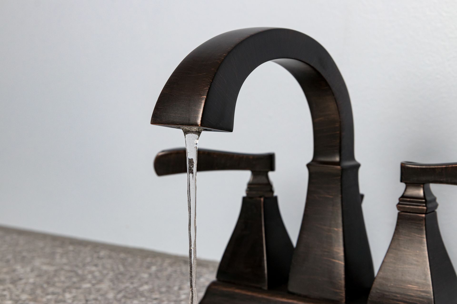 A dark, bronze-colored bathroom faucet with two handles, running a thin stream of water against a light grey wall.