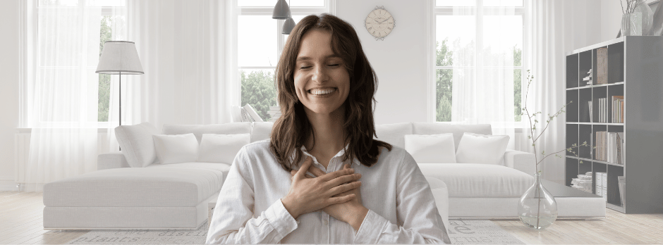 A woman is sitting at a table in a living room with her hands on her chest.