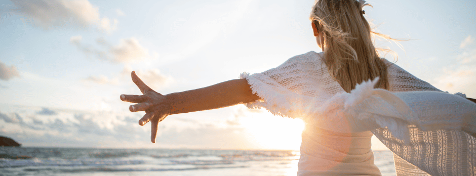 A woman is standing on the beach with her arms outstretched at sunset.
