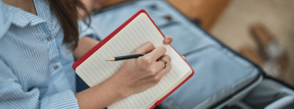A woman is writing in a notebook next to a suitcase.