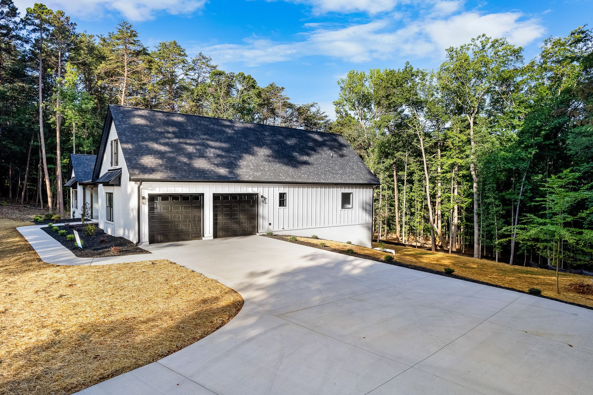 A white house with a black roof is surrounded by trees and a driveway.