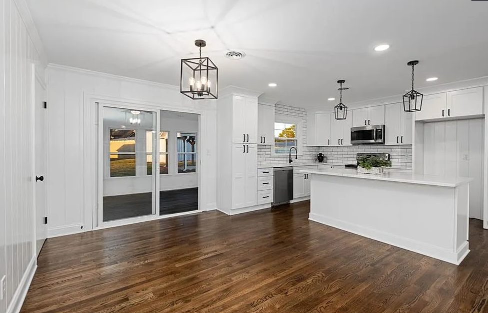 A living room with hardwood floors and a kitchen with white cabinets and stainless steel appliances.
