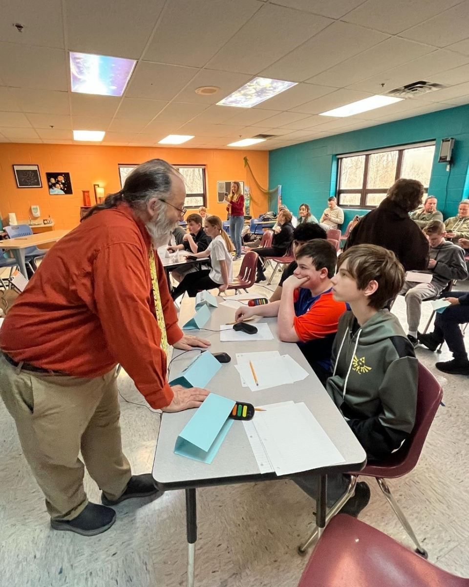 a man is standing in front of a group of children sitting at tables .