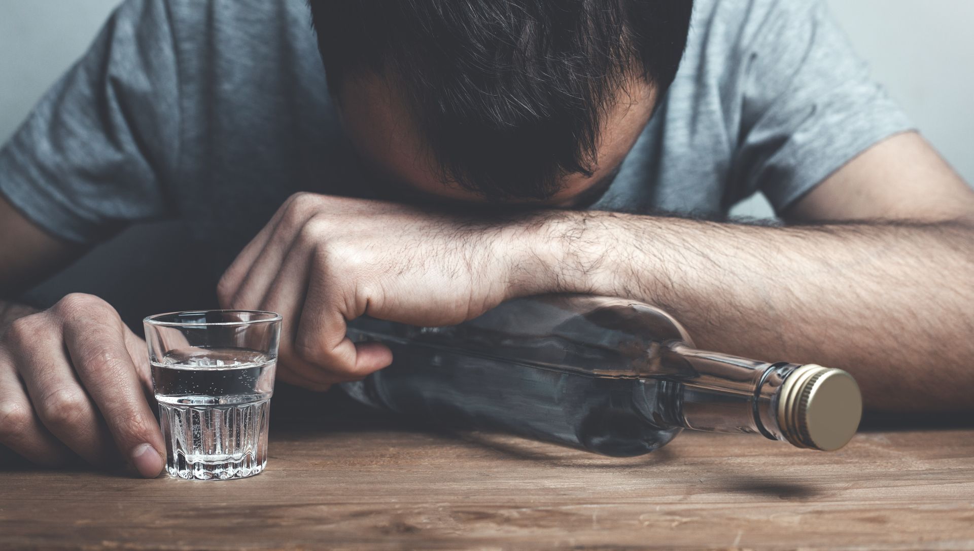 Man with head on table next to empty alcohol bottle and a shot glass.