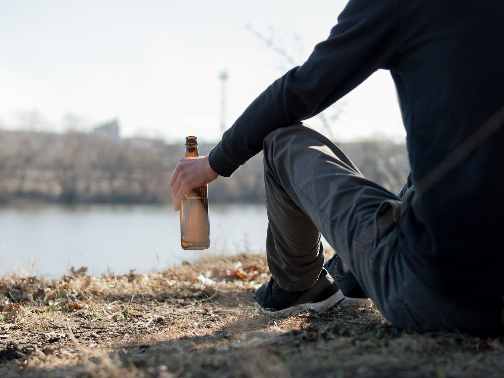 Person sitting by water, holding a beer bottle. Outdoors, sunlight, blurred background.