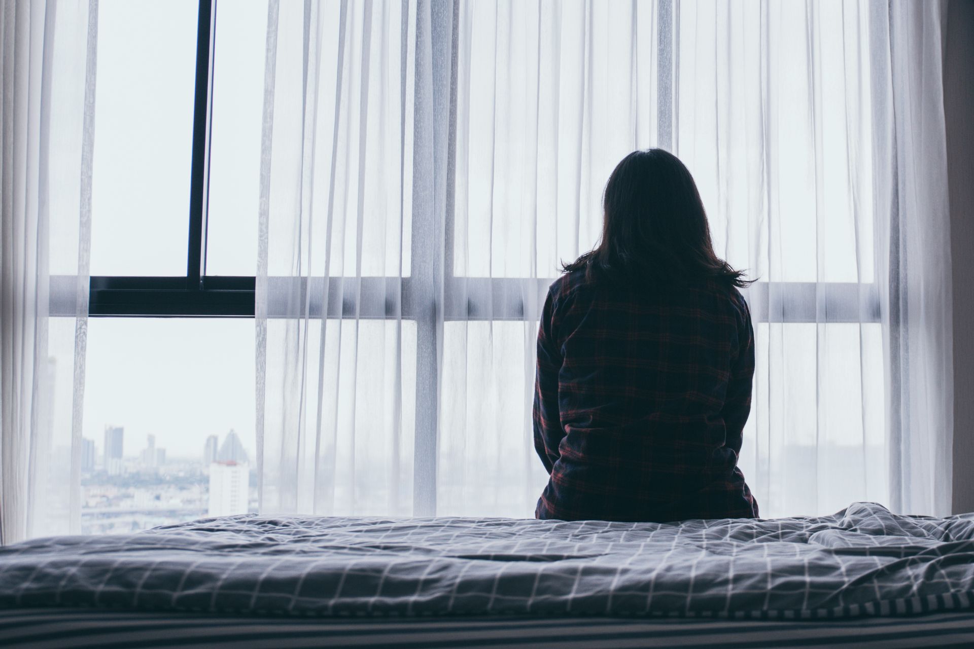 Woman sitting on a bed, looking out a large window with white sheer curtains; city view outside.