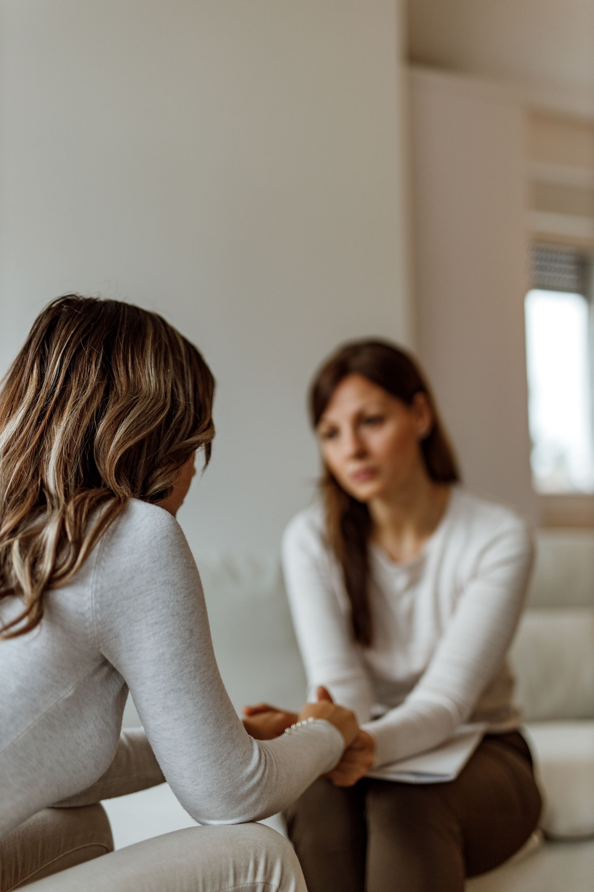 Woman comforting another woman in a light-filled room; one woman holds the other's hands, expressing concern.