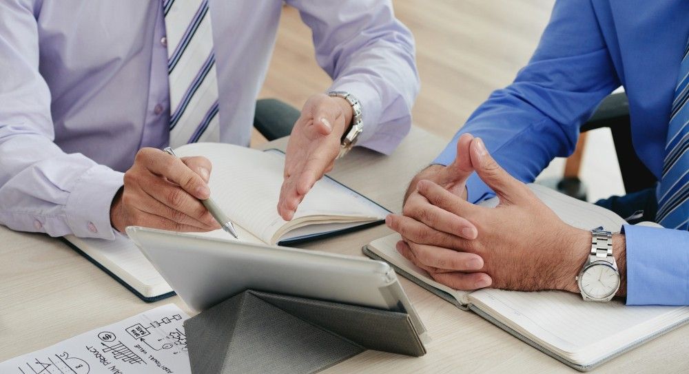 Dos hombres con camisa y corbata discutiendo, usando cuadernos y tabletas en una mesa.