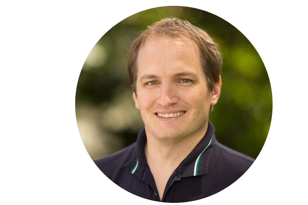 Hombre sonriente con un polo azul marino, al aire libre con fondo verde.