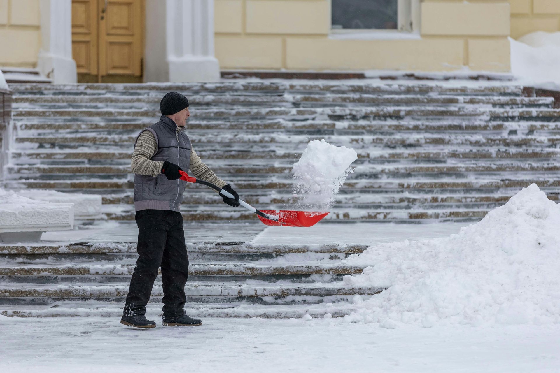 Man shoveling snow from steps of building with red shovel.