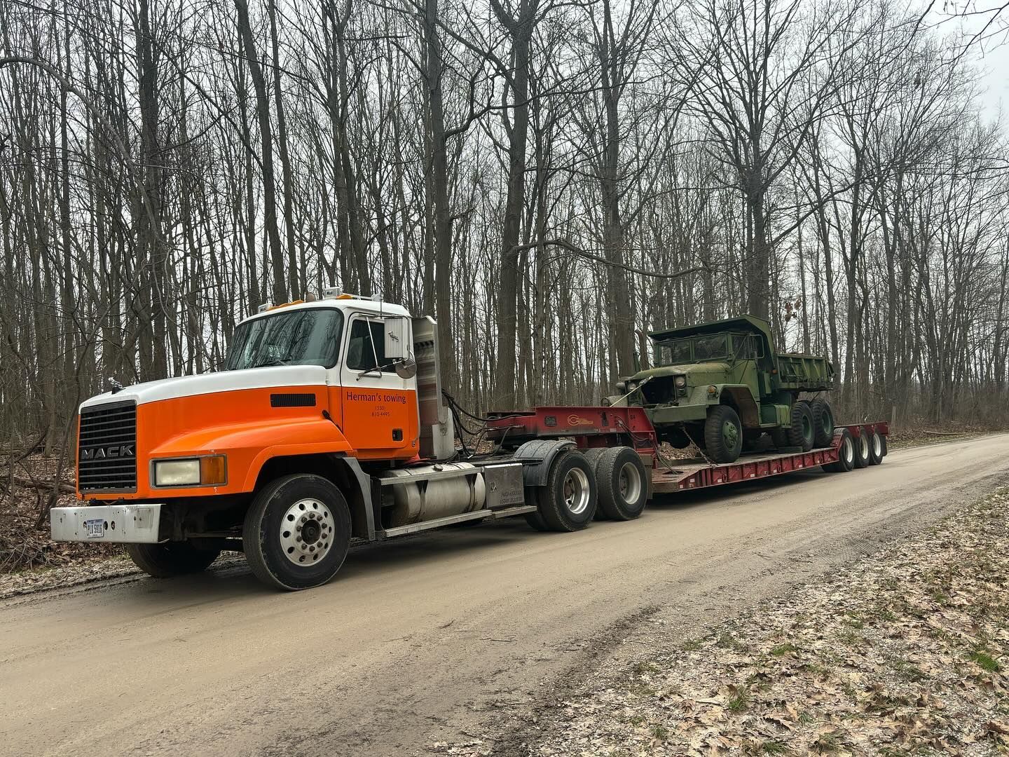 A semi truck is carrying two military vehicles down a dirt road.