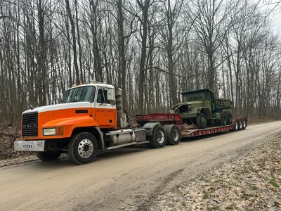 A semi truck is carrying two military vehicles down a dirt road.