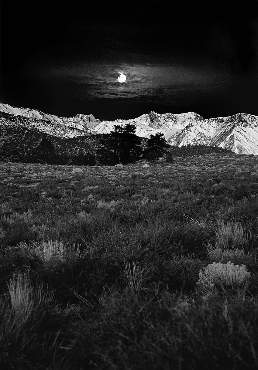 A black and white photo of a full moon rising over a mountain range.