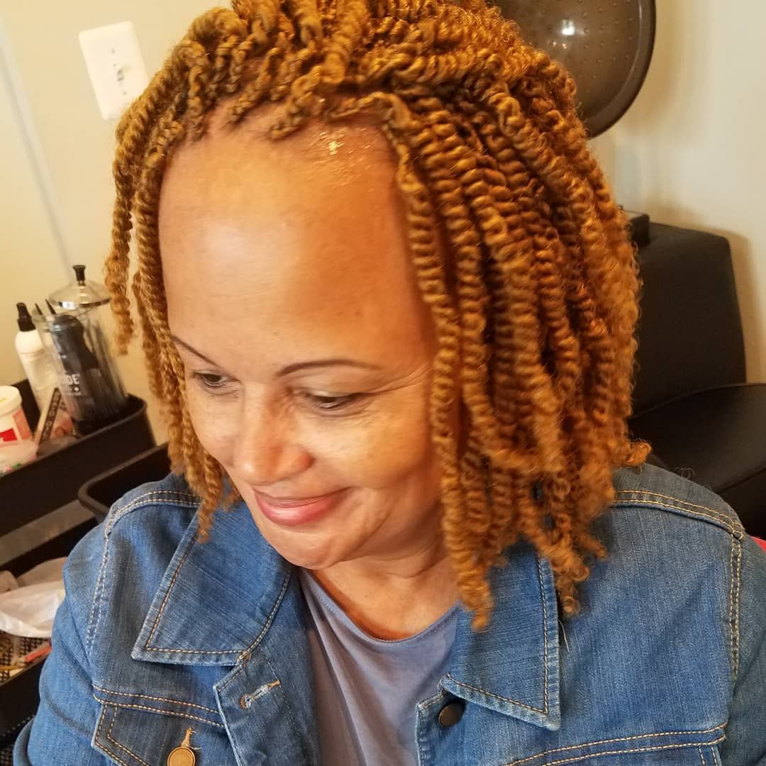 Woman with blonde twist braids smiles, wearing denim jacket, in salon setting.