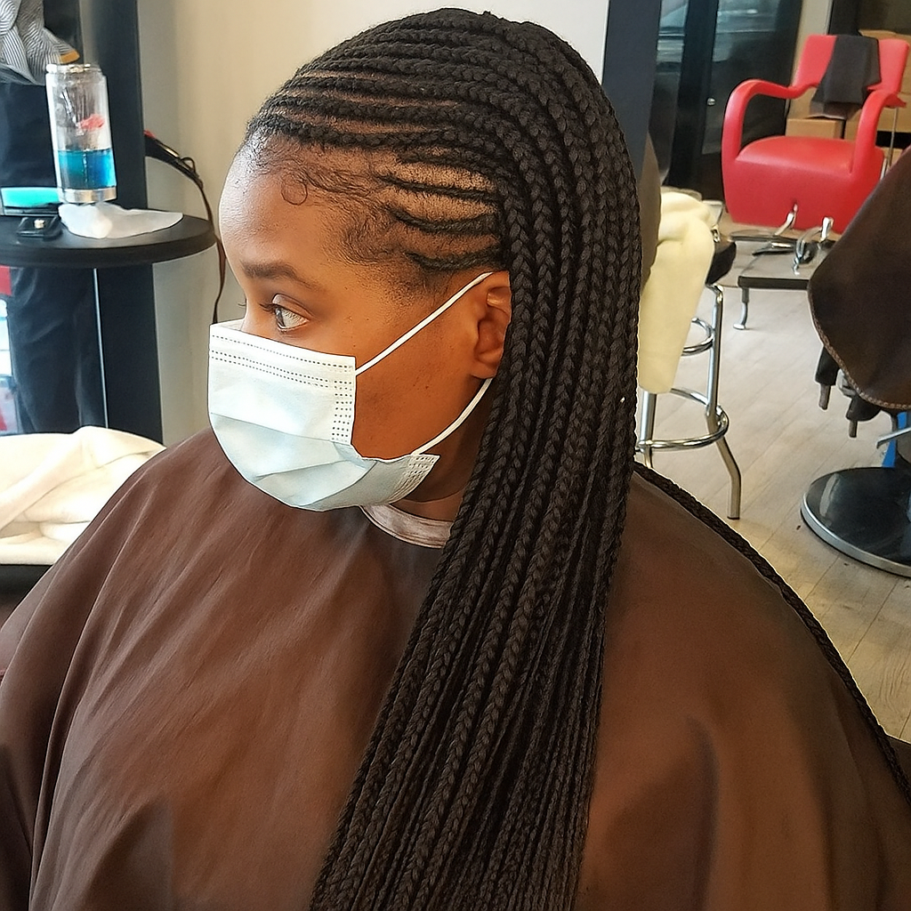 Woman wearing a face mask with braided hair, brown cape, seated in a salon.
