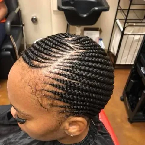 Close-up of a person's head with neatly braided cornrows. The braids are black, angled, and styled with a side part.