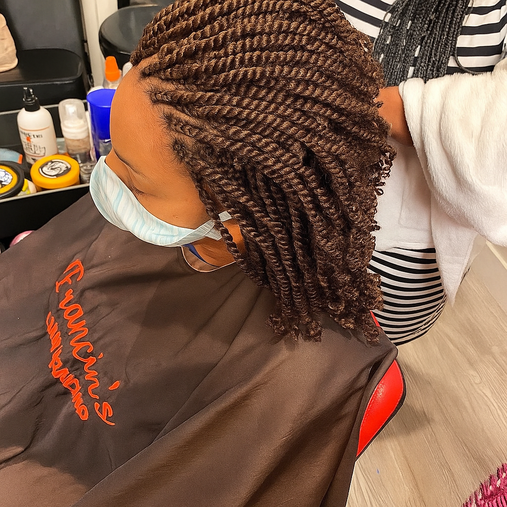 Person with brown twist braids at a salon wearing a face mask and cape; near beauty products.