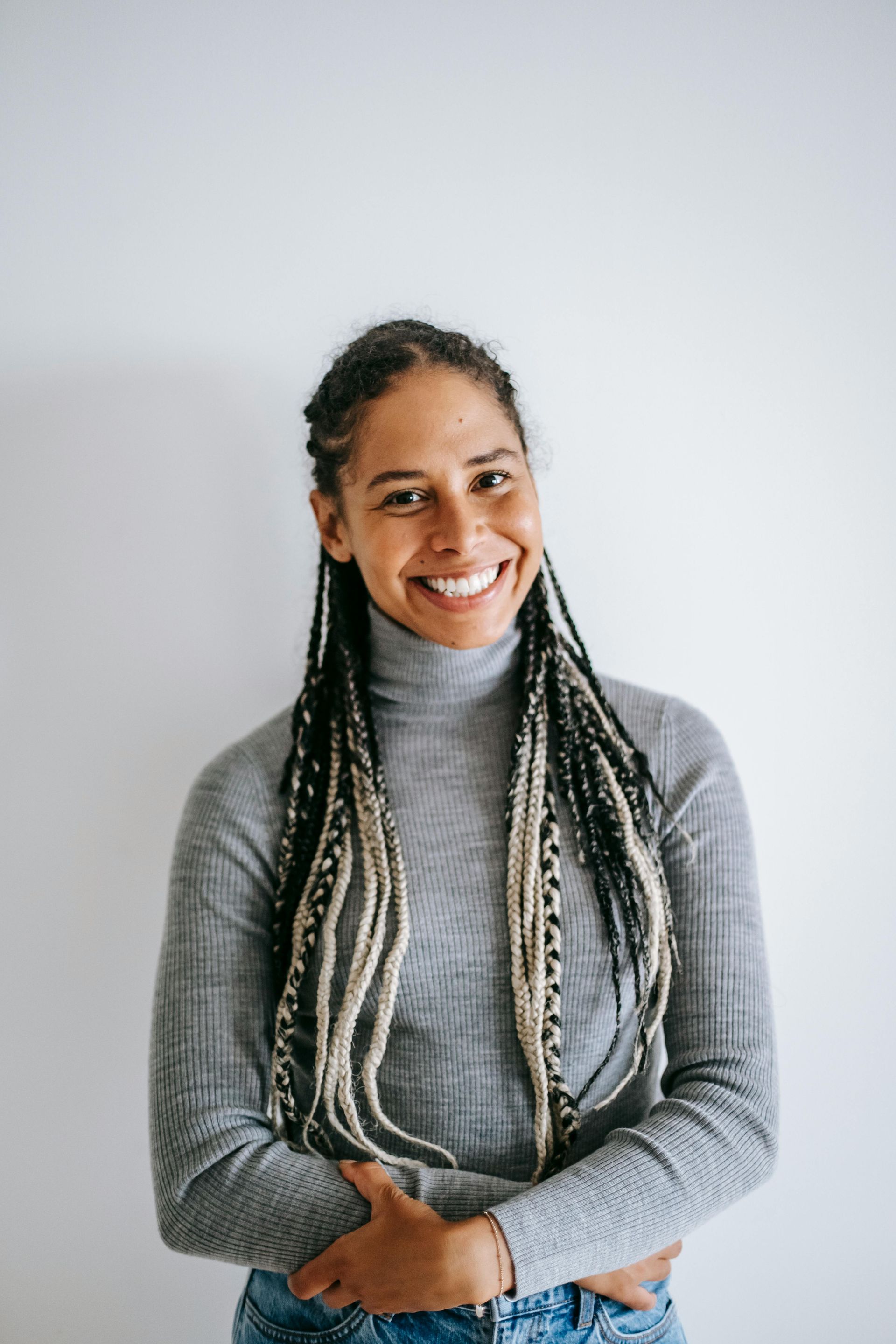 Woman with braided hair smiling, wearing a gray turtleneck sweater and jeans, standing against a white wall.