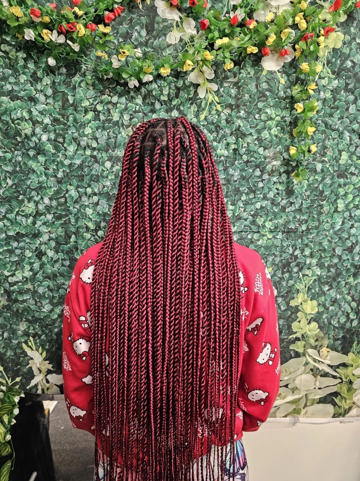 Woman with long, red box braids stands in front of a green wall with flowers. She wears a red sweater.