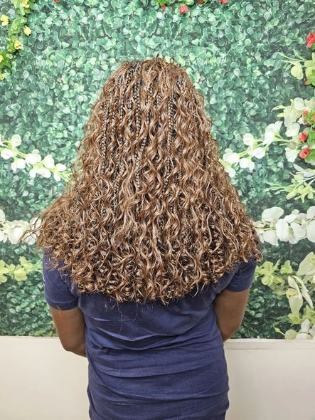 Woman with long, curly, light brown hair wearing a blue shirt, standing in front of a green leafy wall.