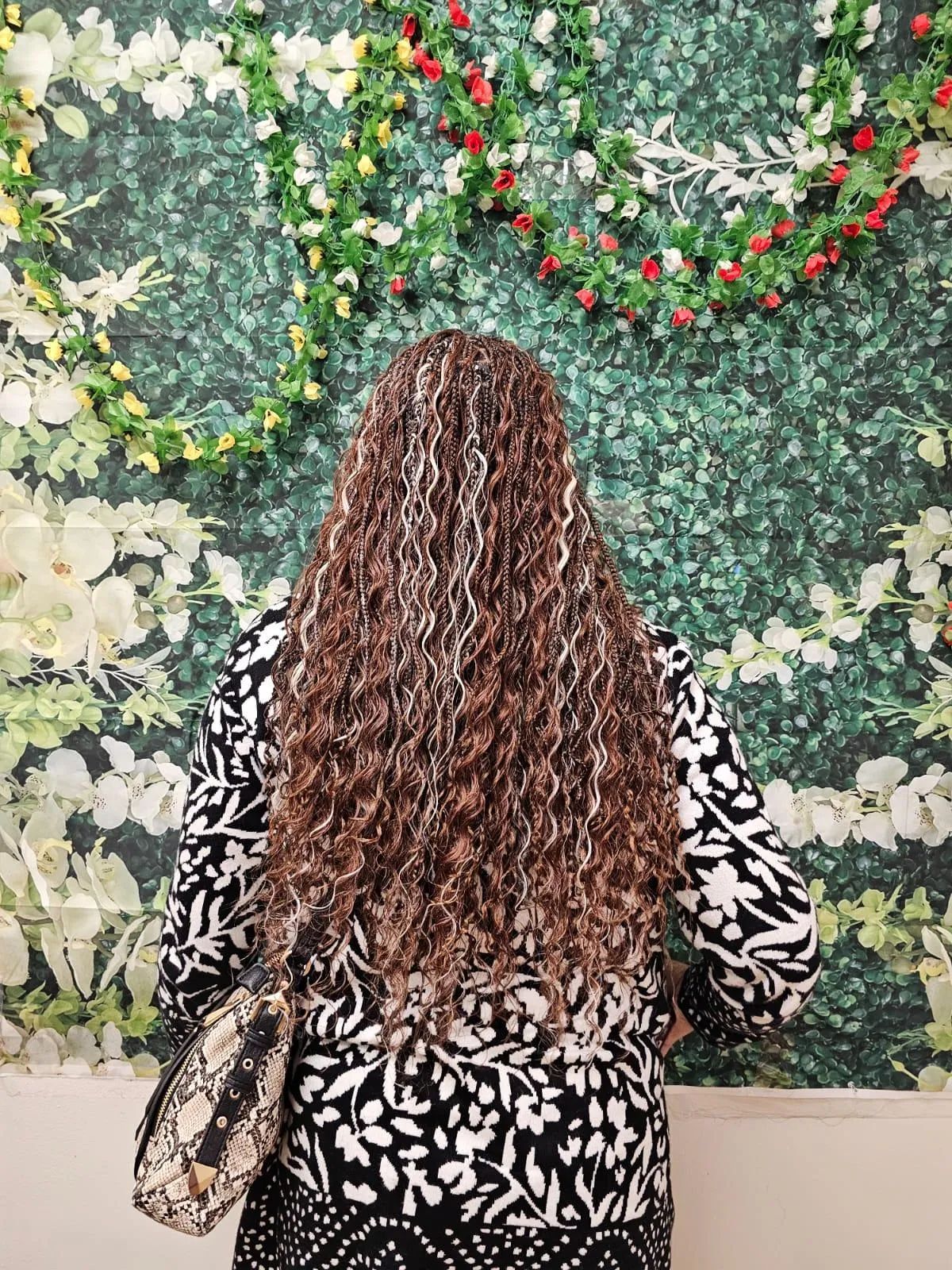 Person with long, curly hair, in black and white patterned top, standing against a floral backdrop.