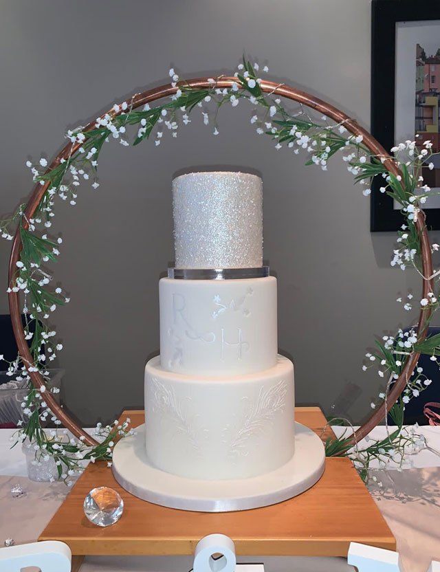 A tall 3-tier white wedding cake, decorated with edible silver glitter all over the top tier, and edible silver spray paint decorating the sides of the other 2 tiers.