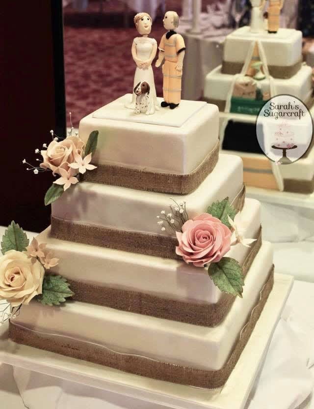 A bespoke 4-tier square wedding cake with 3 figures on top - a lady in a wedding dress, a man in hi-vis clothing, and a dog. The cake is very detailed and very beautiful.