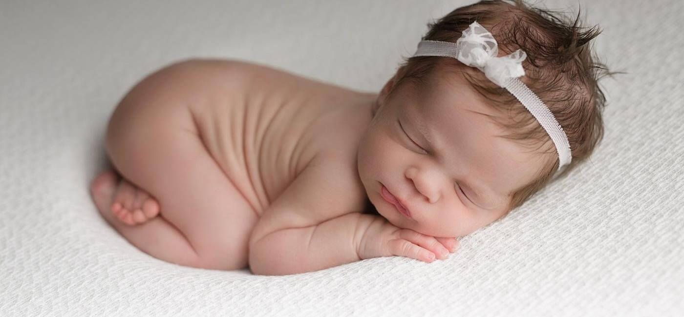 A newborn baby girl wearing a white headband is sleeping on a white blanket.